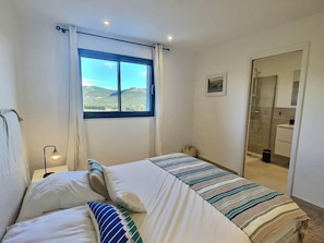 A bedroom with a bed, a bedside lamp, a window with a view of mountains, and a bathroom with a shower.