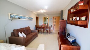 Una sala de estar con un sofá marrón, una mesa de comedor con sillas, un televisor y un aparador de madera con estantes.