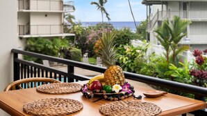 Une table en bois, agrémentée de sets de table en fibres tressées, un ananas et des fleurs tropicales, sur un balcon donnant sur un jardin luxuriant et offrant une vue sur l’océan.