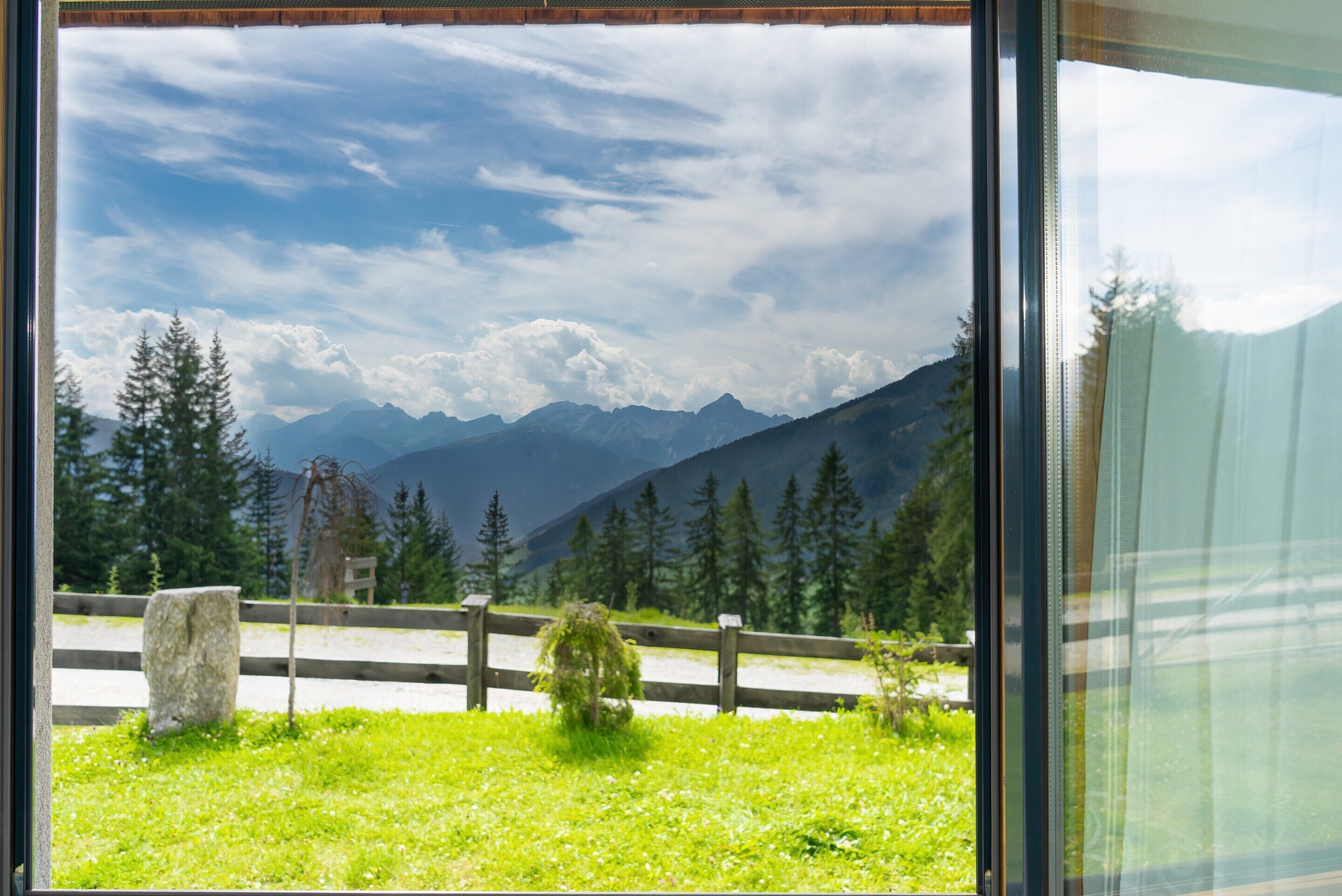 A mountain landscape through a window with a wooden fence and a stone in the foreground.