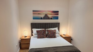 A neatly made bed with a dark headboard, two bedside tables with illuminated lamps, and a colorful abstract pillow. - Lux Modern Apartment in Nuneaton (Nuneaton)