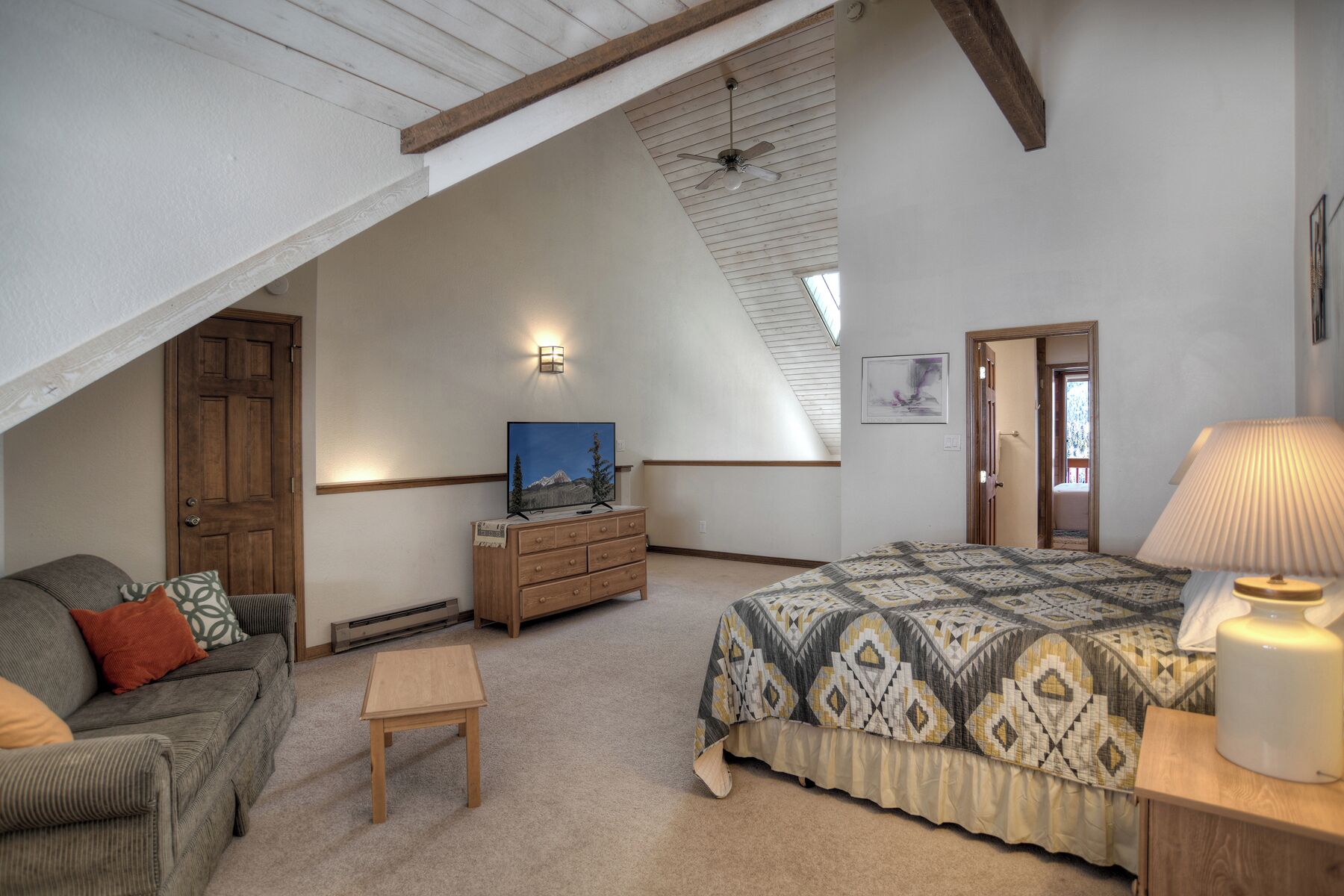 A bedroom with a sloped ceiling, a bed with a patterned bedspread, a wooden dresser, a television, and a lamp.