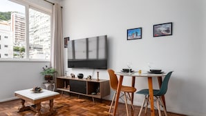 A modern living room with a large TV, wooden floor, a dining table with chairs, and a small table with a bowl of fruit.