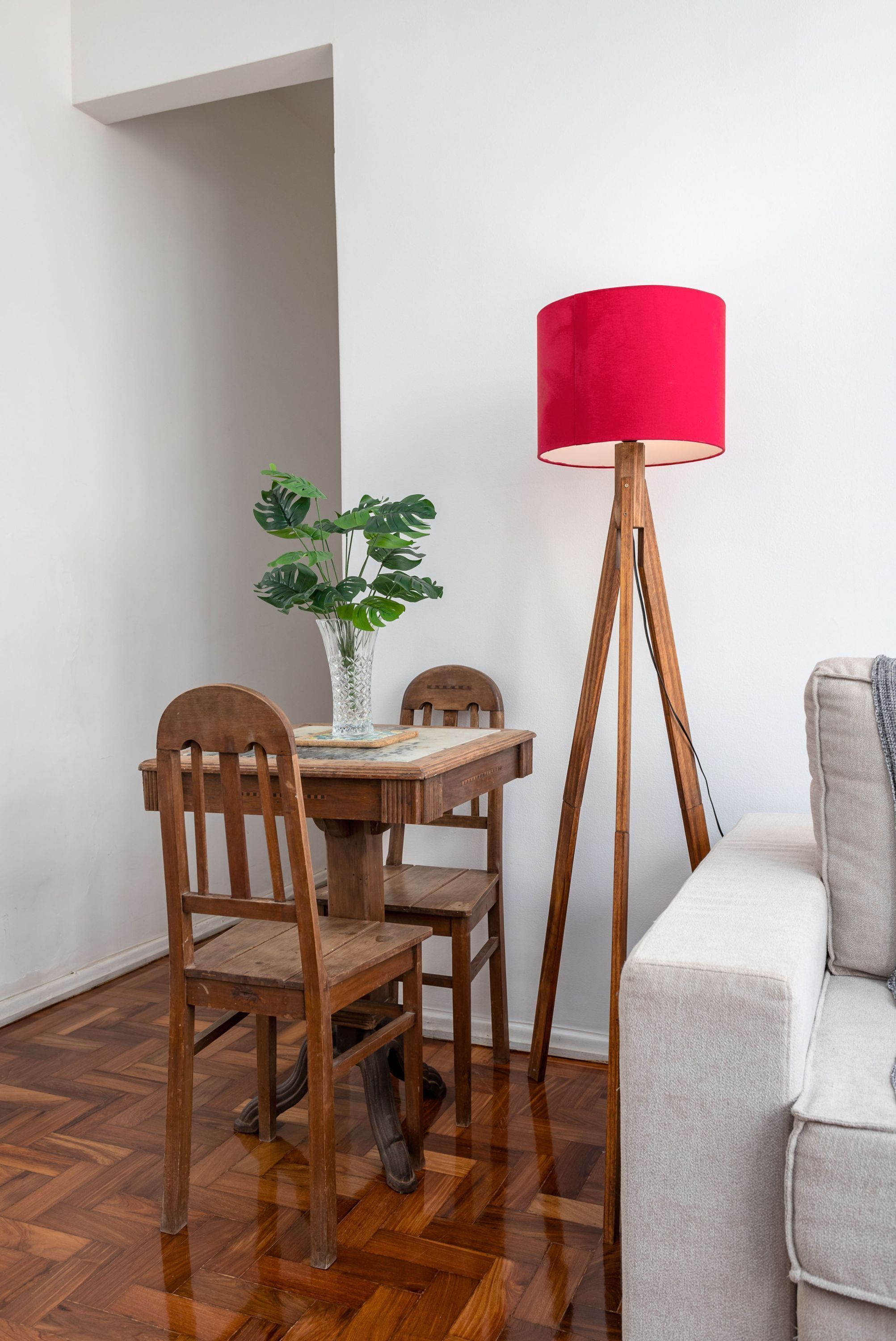 A dining area with a wooden table, chairs, a vase with green plants, and a floor lamp with a red shade.