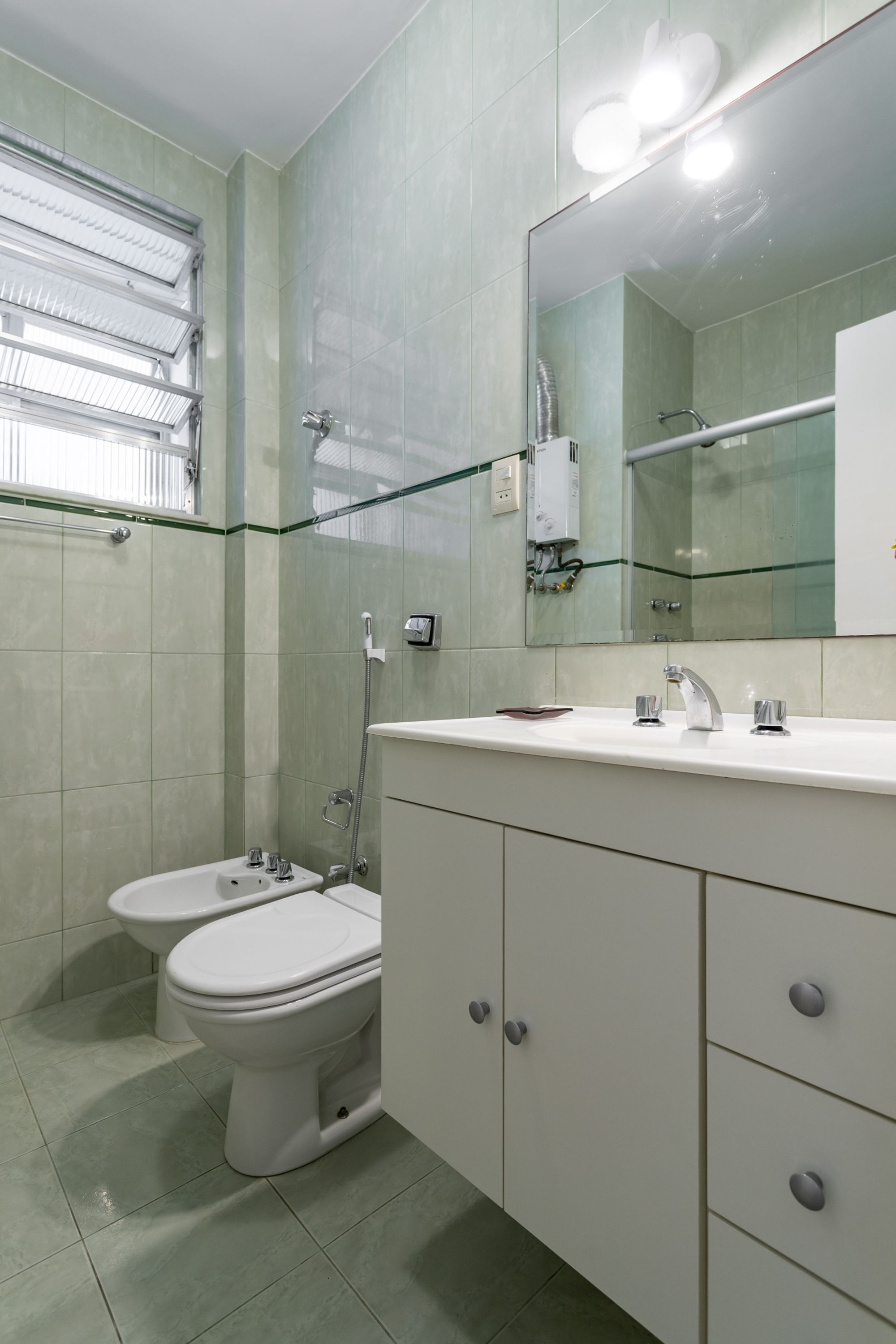 Un baño con un lavamanos blanco, dos inodoros, una cabina de ducha con mampara de vidrio y un espejo con iluminación.