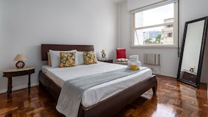 A bedroom with a wooden bed, a nightstand, a lamp, a chair, a window with a view of buildings, and a mirror.