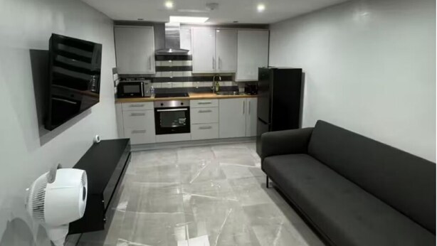 A modern kitchen with a built-in oven, microwave, and stainless steel range hood. There is a black sofa and a white fan in the living area. - Place to stay in Erith (Erith)