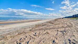 Une plage de sable avec des empreintes de pas et des traces de pneus, un ciel bleu clair et des bâtiments qui se dessinent à l’horizon.