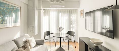 Una sala moderna con un sofá gris, una mesa de comedor redonda con sillas, un mueble negro para televisor y un ventanal con cortinas traslúcidas.
