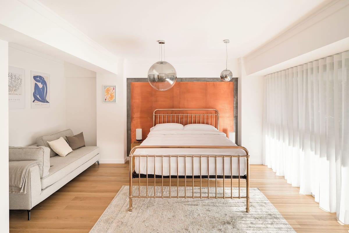 A modern bedroom with a metal bed frame, a grey sofa, and a large window with sheer curtains.