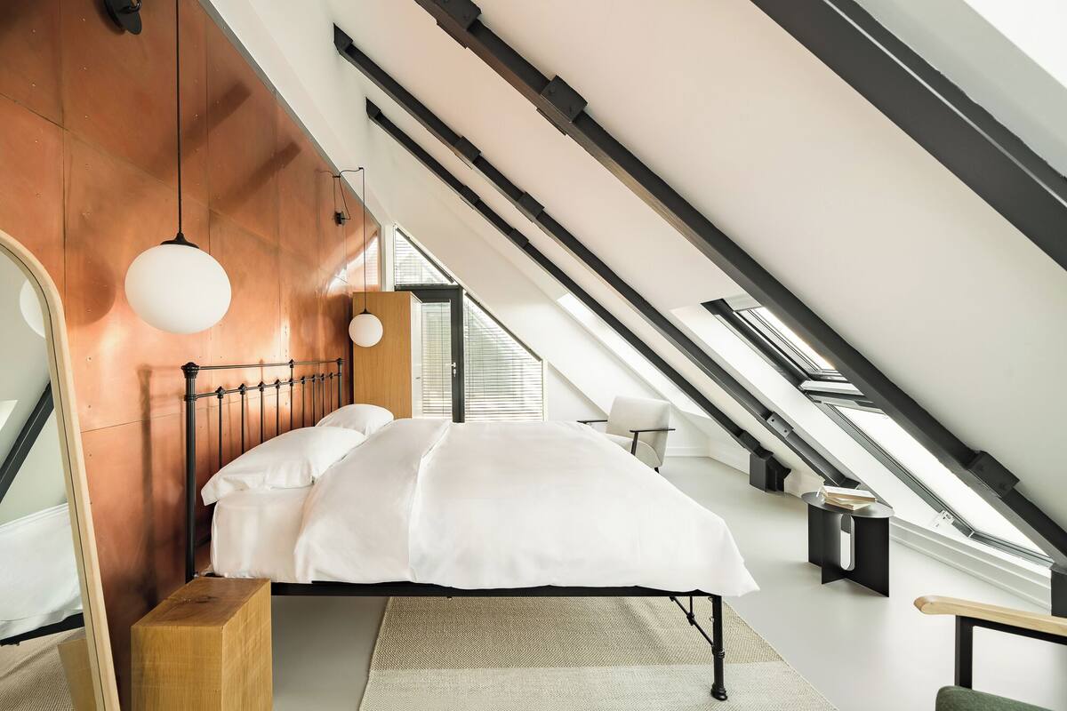 A modern loft bedroom with a sloped ceiling, exposed beams, a large bed, a hanging light fixture, and a view of the sky through a skylight.