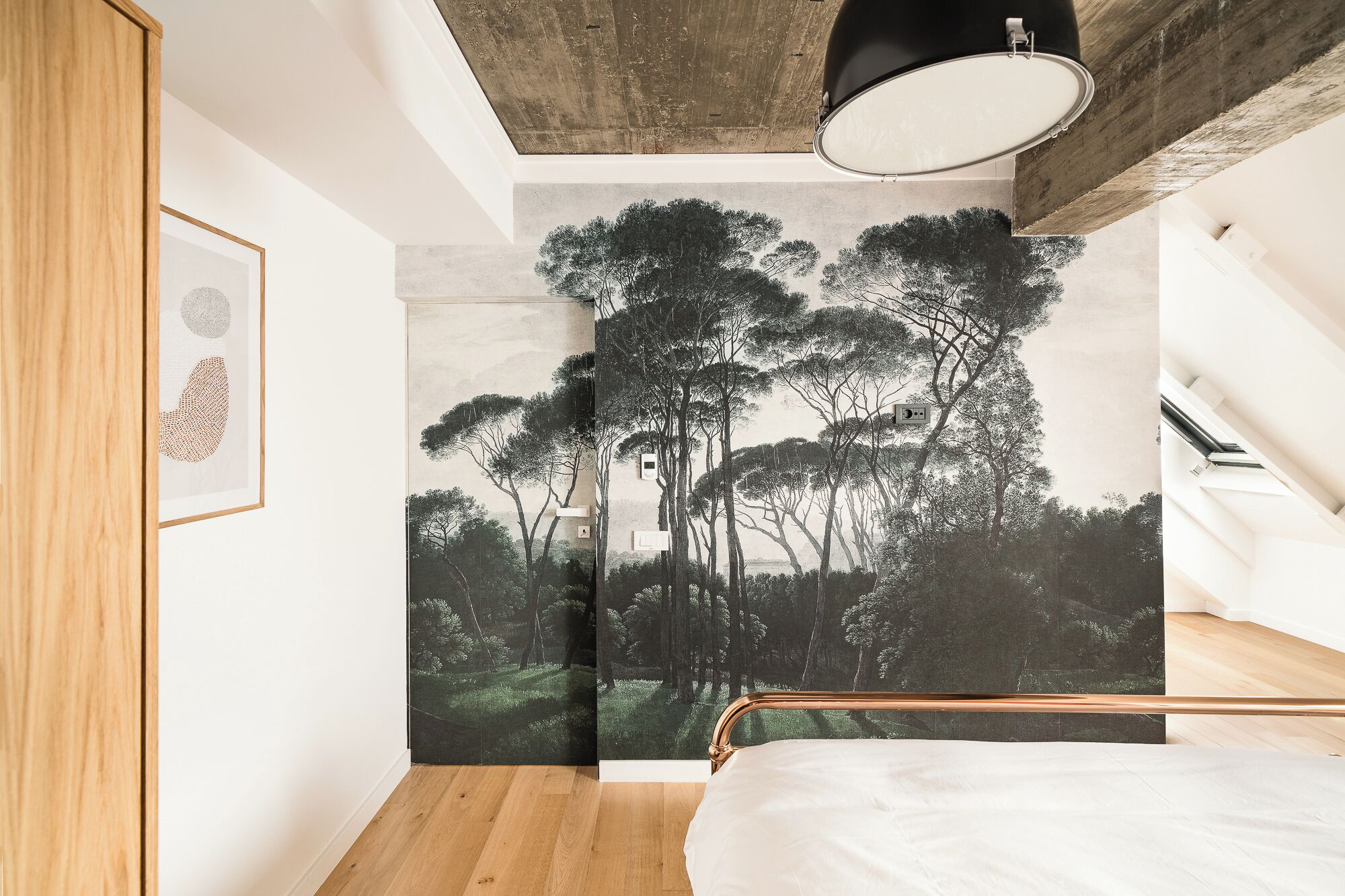 A bedroom with a bed, a wooden wardrobe, a ceiling light, and a wall mural of trees.