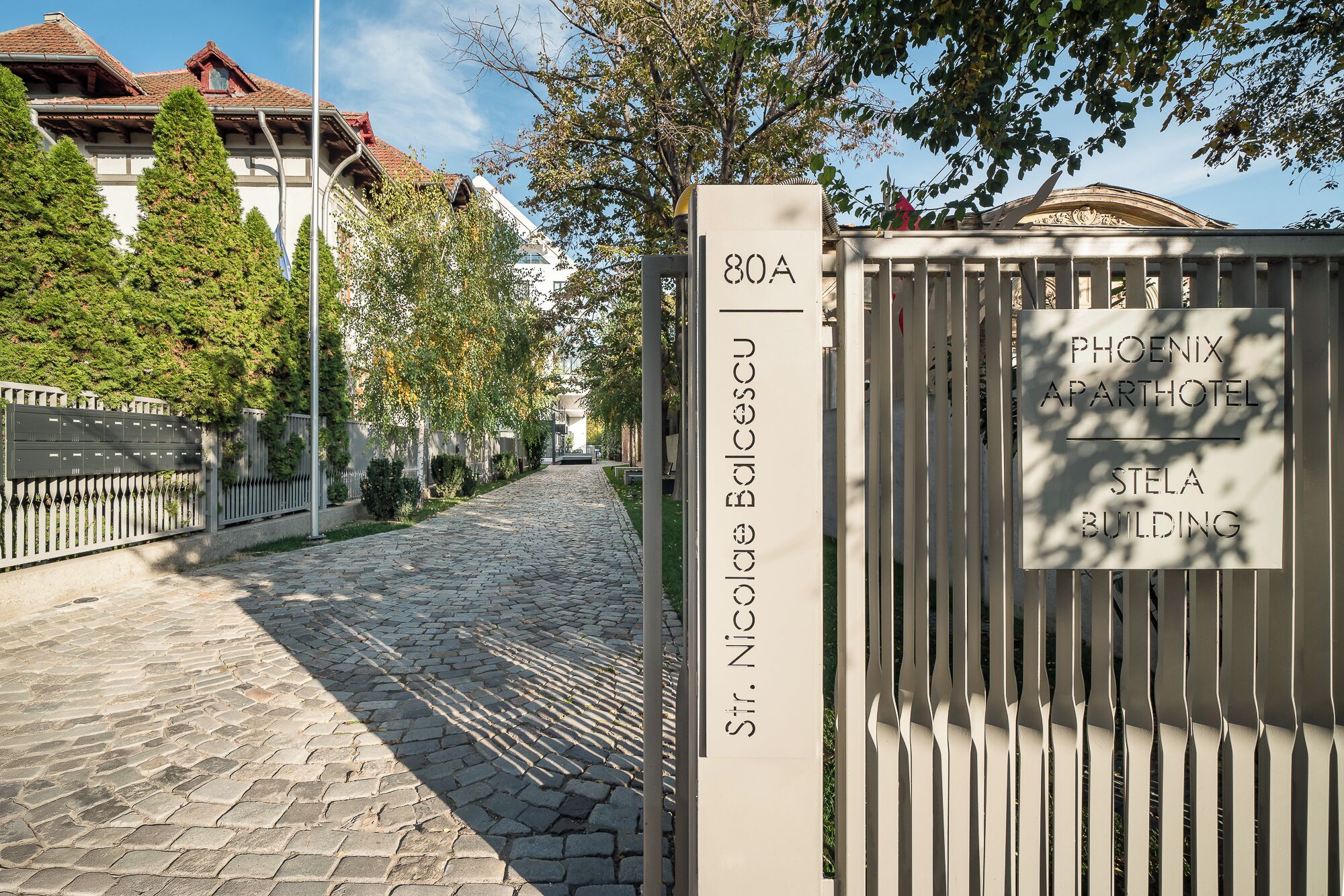 A cobblestone street leading to a gated entrance with a sign for 'St. Nicholas Balczeu' and 'Phoenix Apartotel Stela Building'.