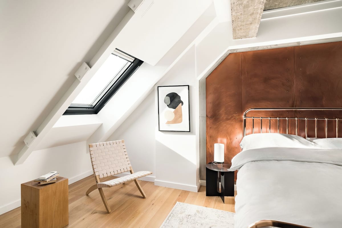 A bedroom with a sloped ceiling, a bed, a wooden chair, a small table with a lamp, and a framed picture on the wall.