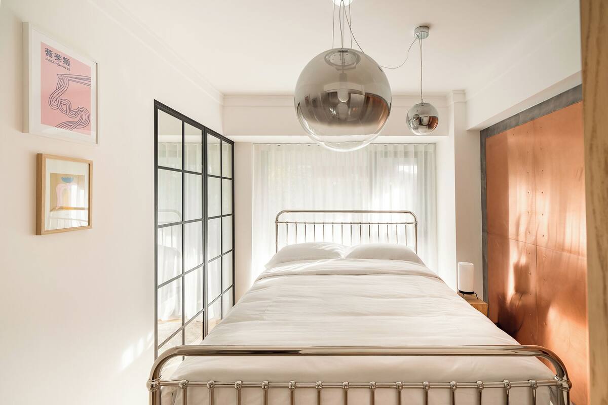 A modern bedroom with a metal bed frame, a large window with blinds, and a wooden door.