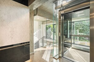 Un ascensor moderno con puertas de cristal, un pilar de hormigón y vistas a una escalera que conduce a un espacio exterior con vegetación.