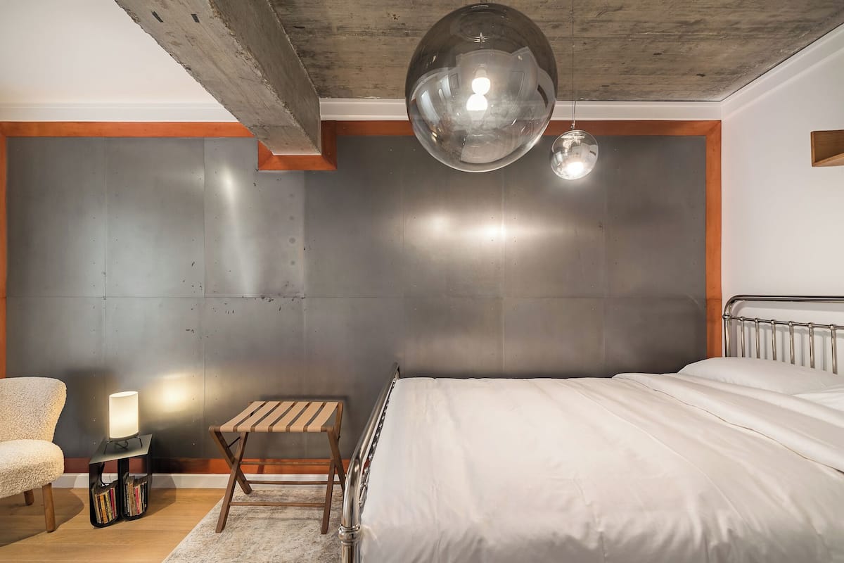 A modern bedroom with a large bed, a small wooden stool, a side table with a lamp, and a large mirror on the wall.