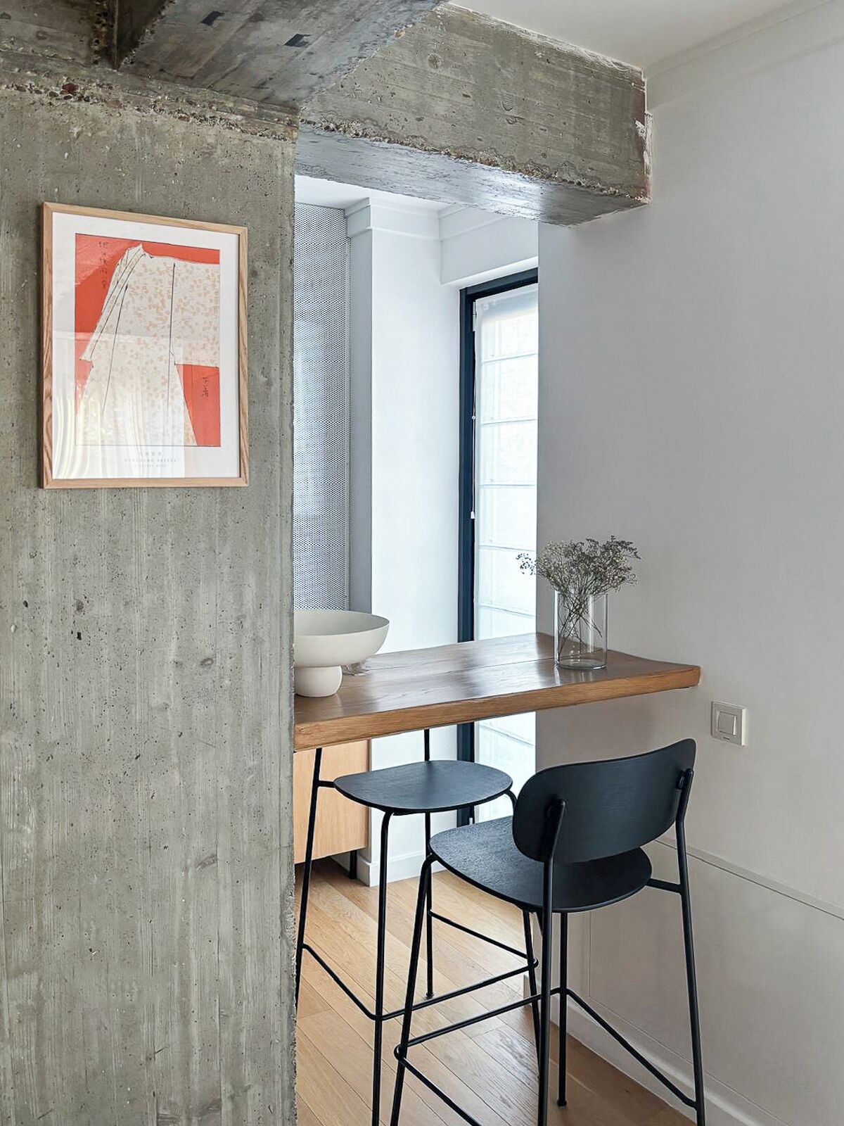 Een keuken met een betonnen pilaar, een houten aanrecht, twee zwarte stoelen en een lijst met een foto aan de muur.