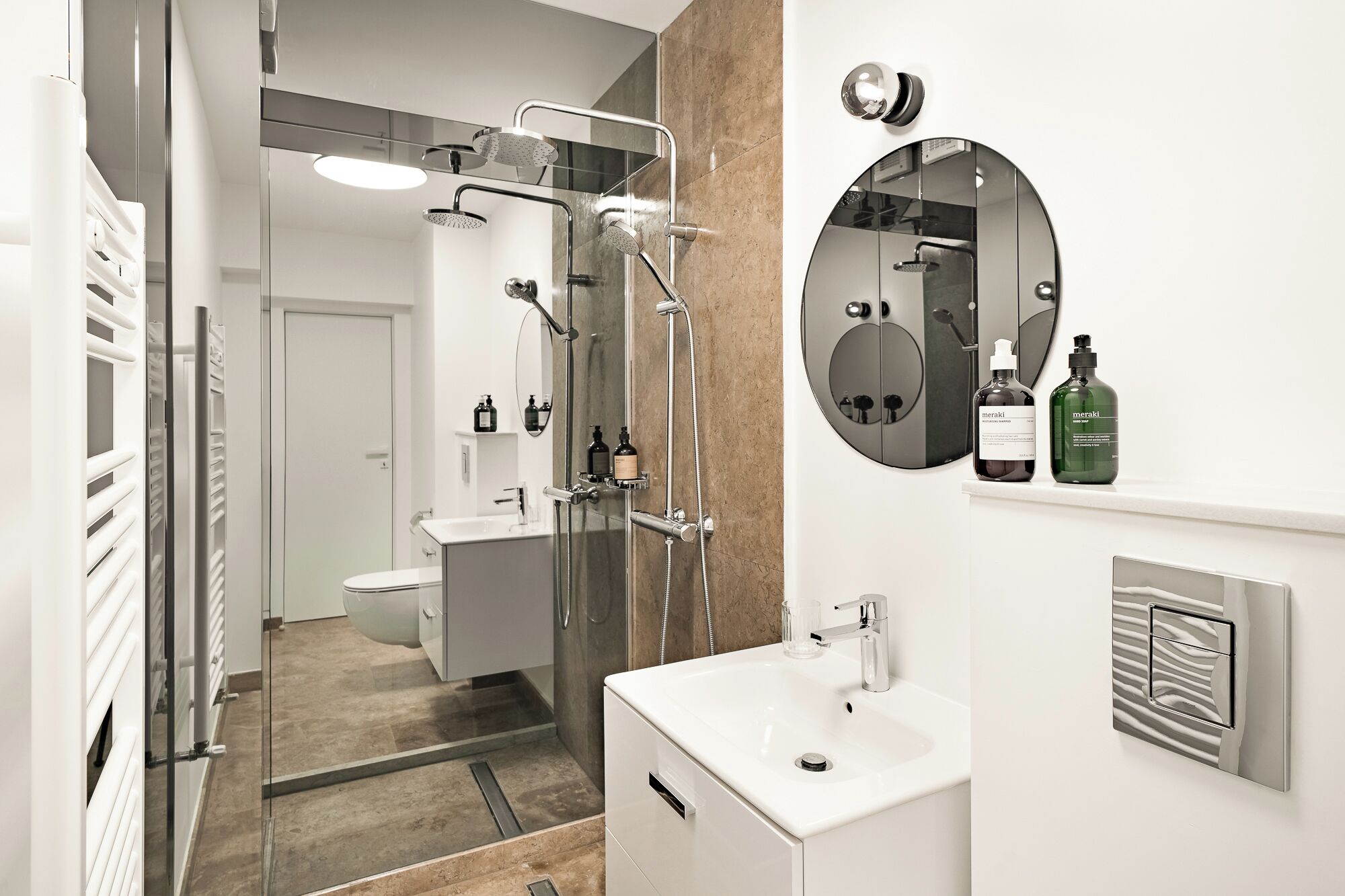 A modern bathroom with a shower, toilet, and sink. There are two bottles of Meraki on the sink.