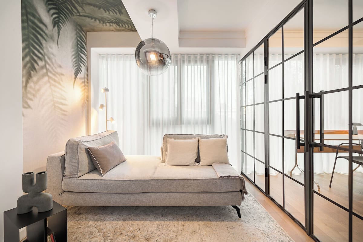 A modern living room with a grey sofa, two cushions, a hanging light fixture, and a large window with sheer curtains.