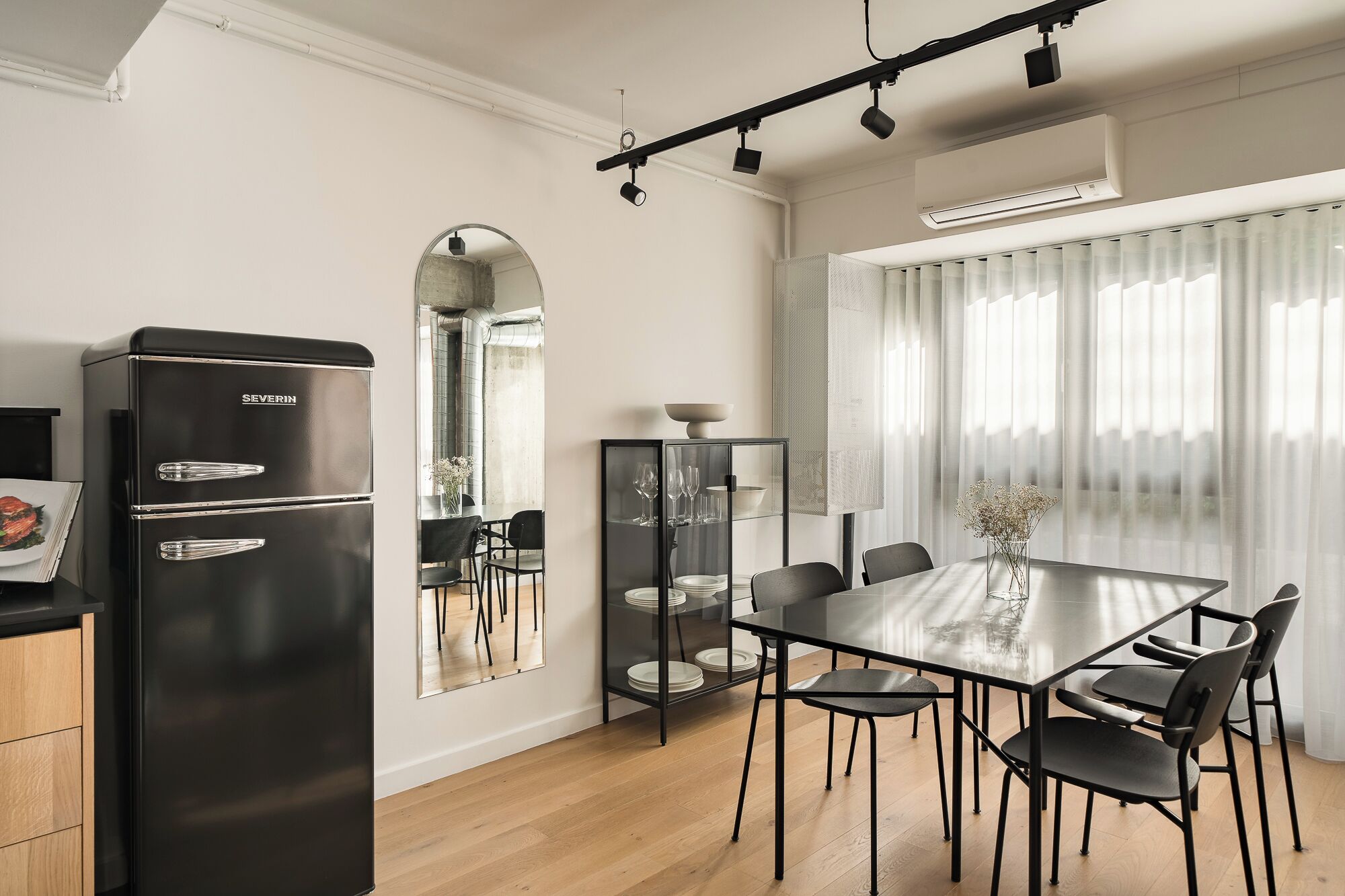 Una cocina moderna con un refrigerador negro, armarios de madera, una mesa de comedor con sillas y un gran ventanal con cortinas traslúcidas.