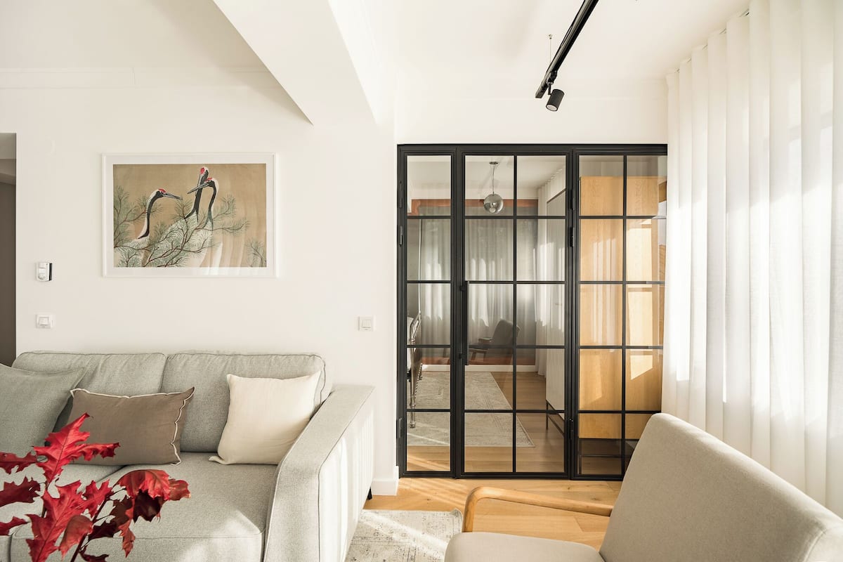 A living room with a grey sofa, a wooden floor, a glass door leading to a dining area, and a framed picture on the wall.