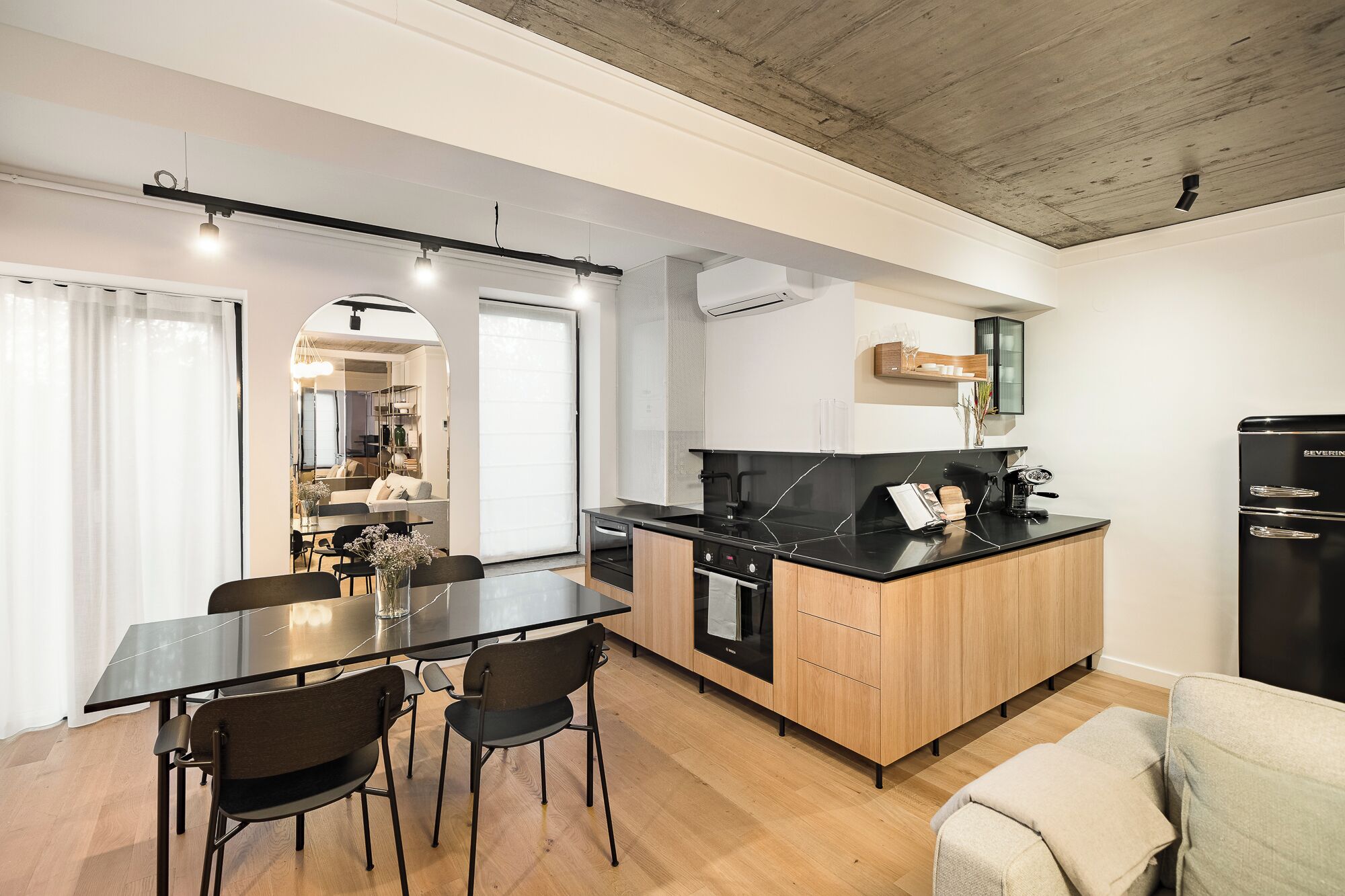 Cocina moderna con encimera negra, armarios de madera y horno integrado. Cuenta con un comedor con mesa y sillas, y un sofá en la esquina.