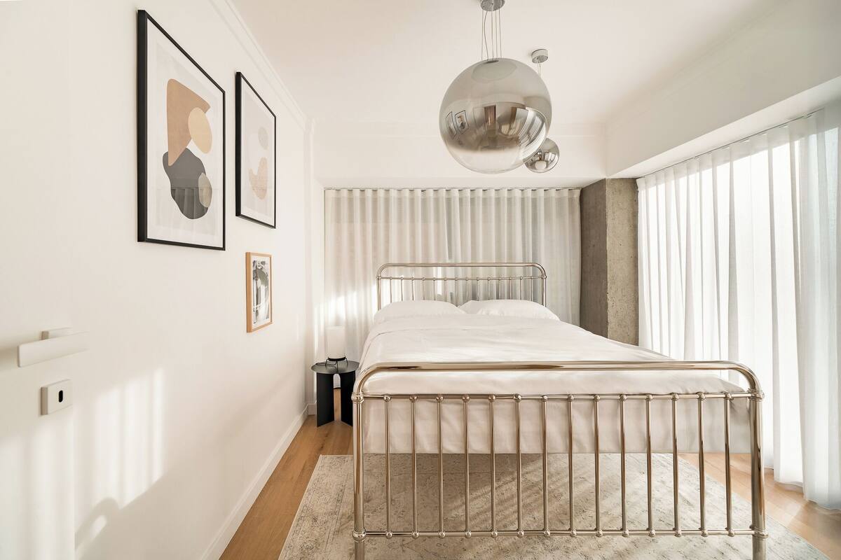 A modern bedroom with a metal bed frame, a large window with sheer curtains, and a hanging spherical light fixture.
