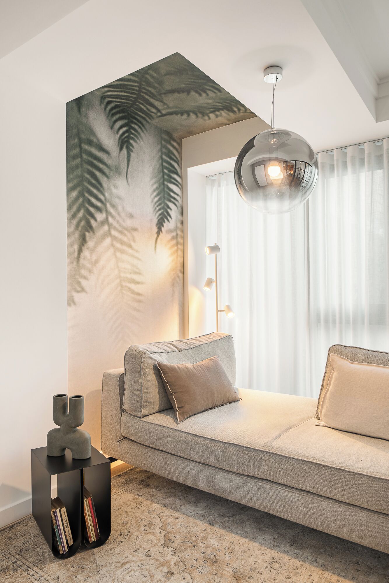 Una sala de estar moderna con un sofá gris, una pared decorativa con motivos de hojas, una mesita auxiliar negra con libros e iluminación colgante.