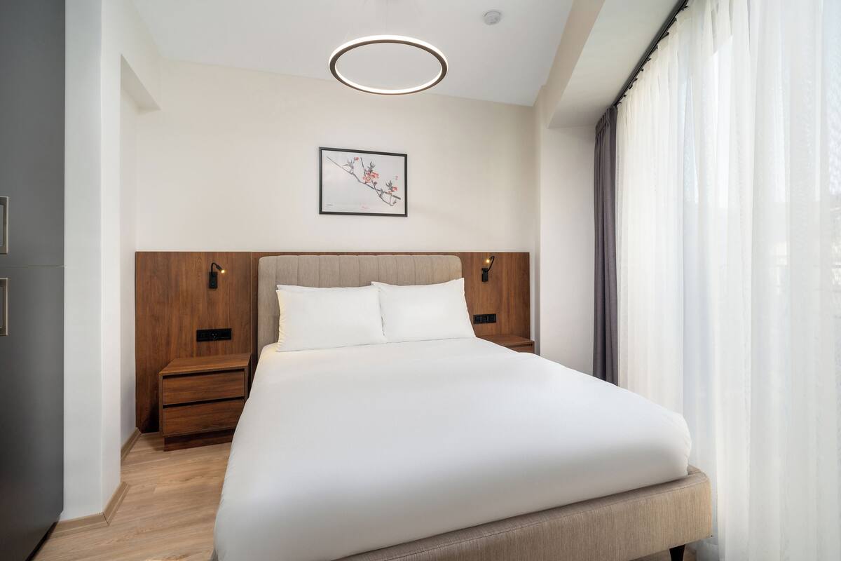 A modern hotel room with a large bed, wooden headboard, bedside tables, and a ceiling light fixture.