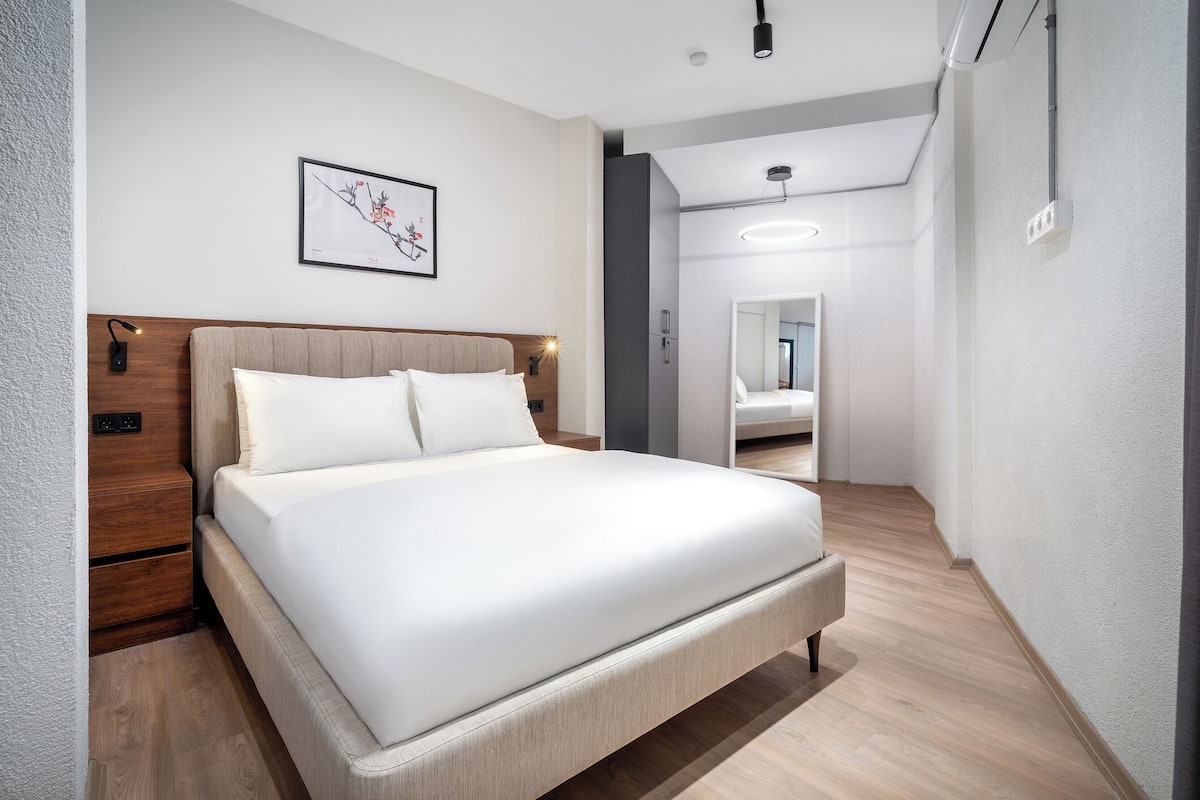 A modern hotel room with a large bed, a wooden headboard, a bedside table, and a nightstand. There is a framed artwork on the wall and a mirror in the room.