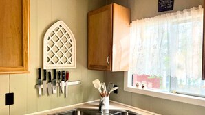 Une cuisine dotée de deux éviers, de meubles de rangement en bois, d’une fenêtre ornée de rideaux en dentelle et d’un porte-couteaux fixé au mur.