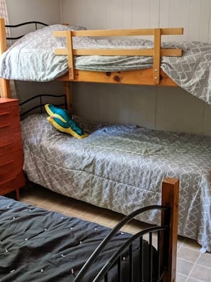 A bunk bed with a wooden frame, a lower bunk with a patterned blanket, and a single bed with a dark blanket.