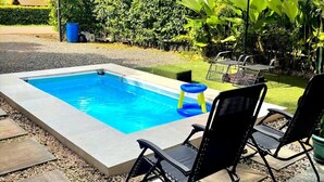 Une piscine rectangulaire à l’intérieur bleu, entourée d’une terrasse carrelée avec des dalles permettant de traverser. On y trouve deux chaises longues noires et une petite table avec un parasol.