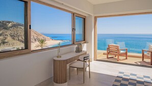 Een moderne kamer met grote ramen met uitzicht op een strand, een bureau met stoel, en een balkon met twee stoelen.