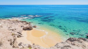 Een strand met helder turquoise water, een zandstrand en rotsformaties.