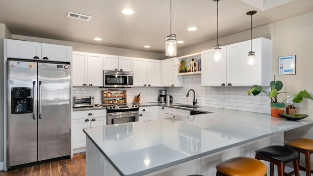 A modern kitchen with white cabinets, stainless steel appliances, a central island with stools, pendant lighting, and a visible vent on the ceiling.