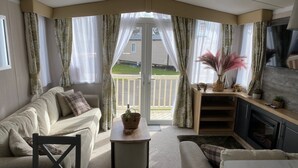 Un salon confortable avec un canapé, une table basse et un téléviseur. Il y a un balcon avec des rideaux et une vue sur une maison.
