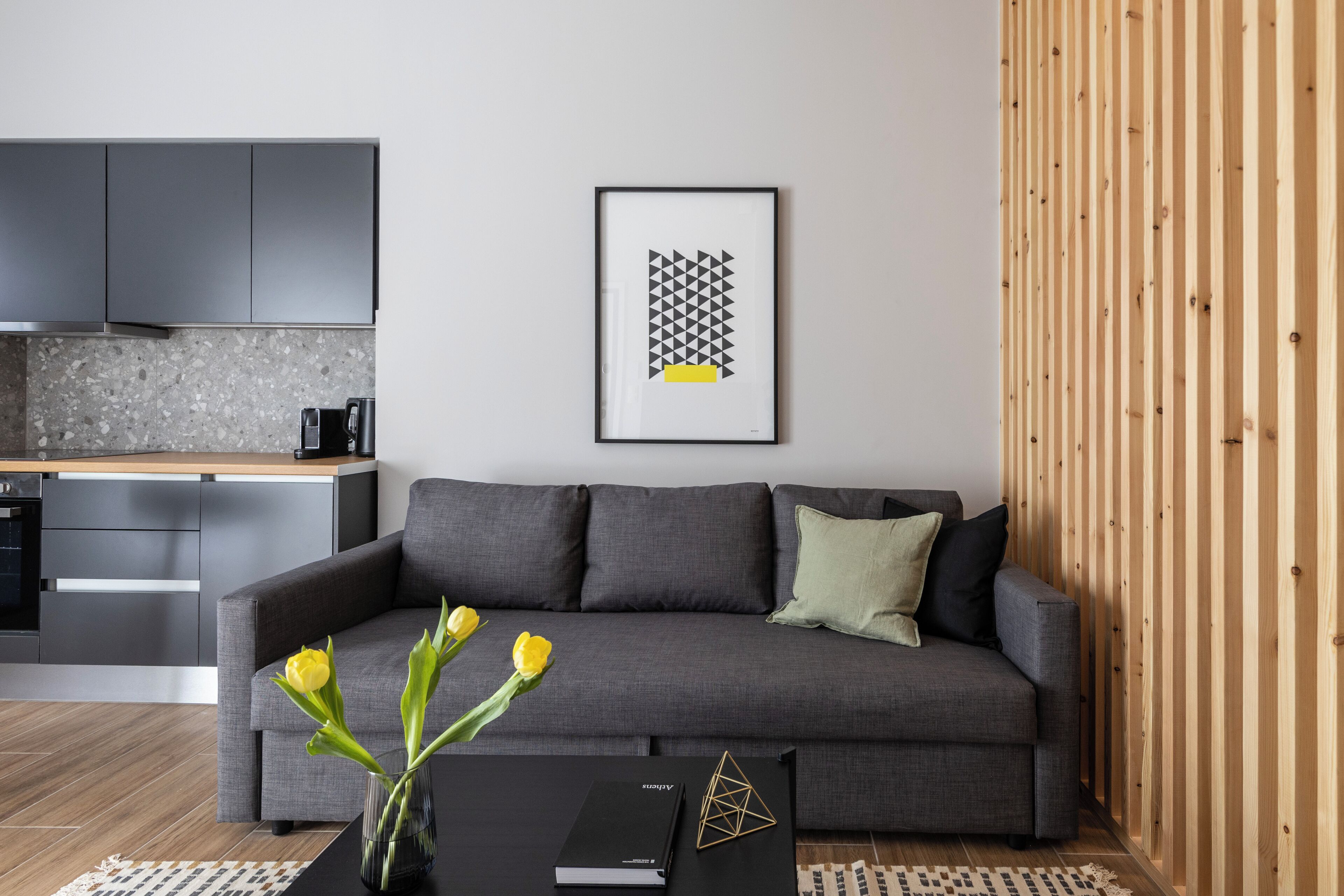 A modern living room with a grey sofa, a coffee table with a book and a geometric decoration, a vase with yellow flowers, and a framed abstract art piece on the wall.