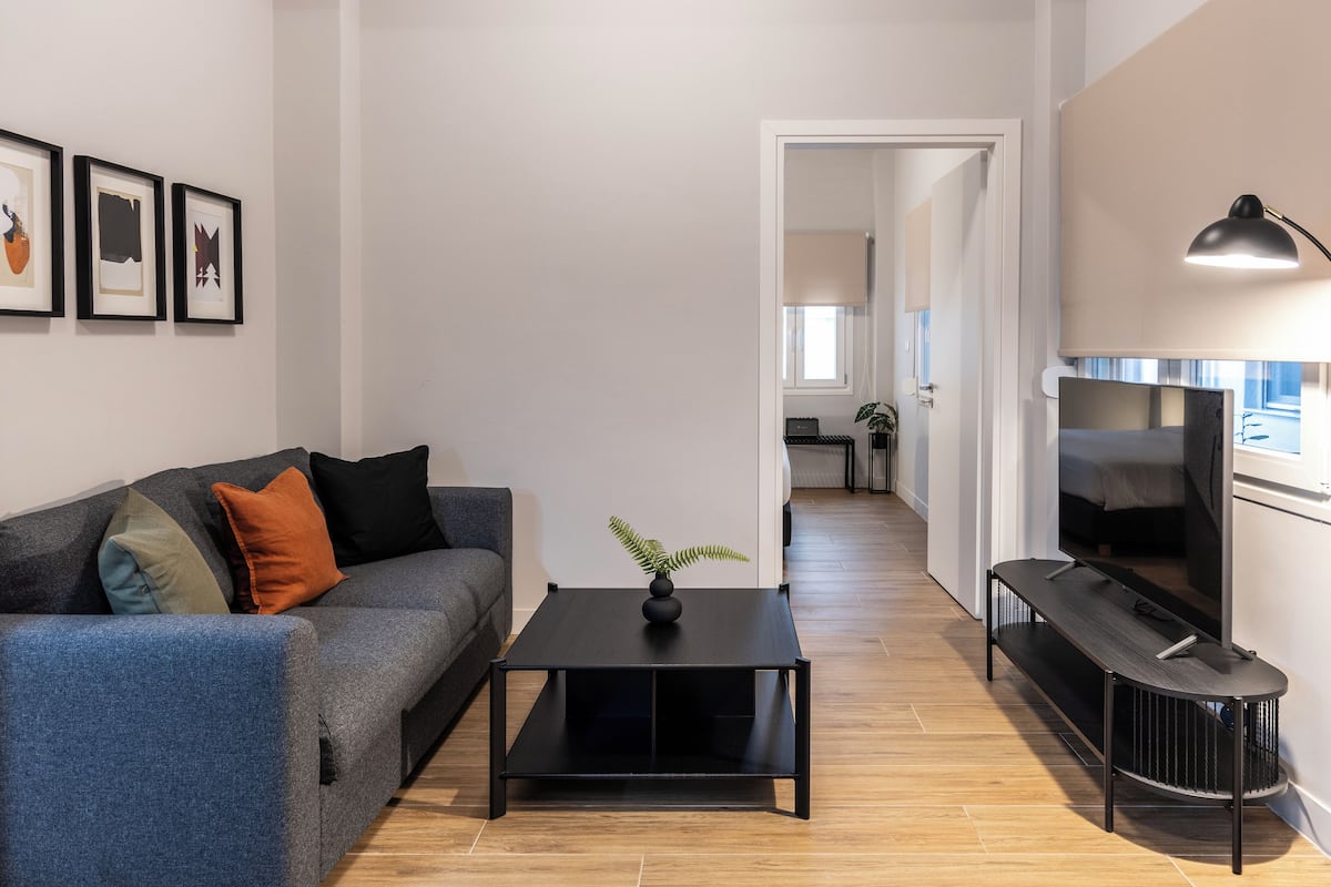 Ein modernes Wohnzimmer mit einer Couch, einem Couchtisch und einem Fernseher. An der Wand hängt ein gerahmtes Bild und auf dem Couchtisch steht eine Pflanze.
