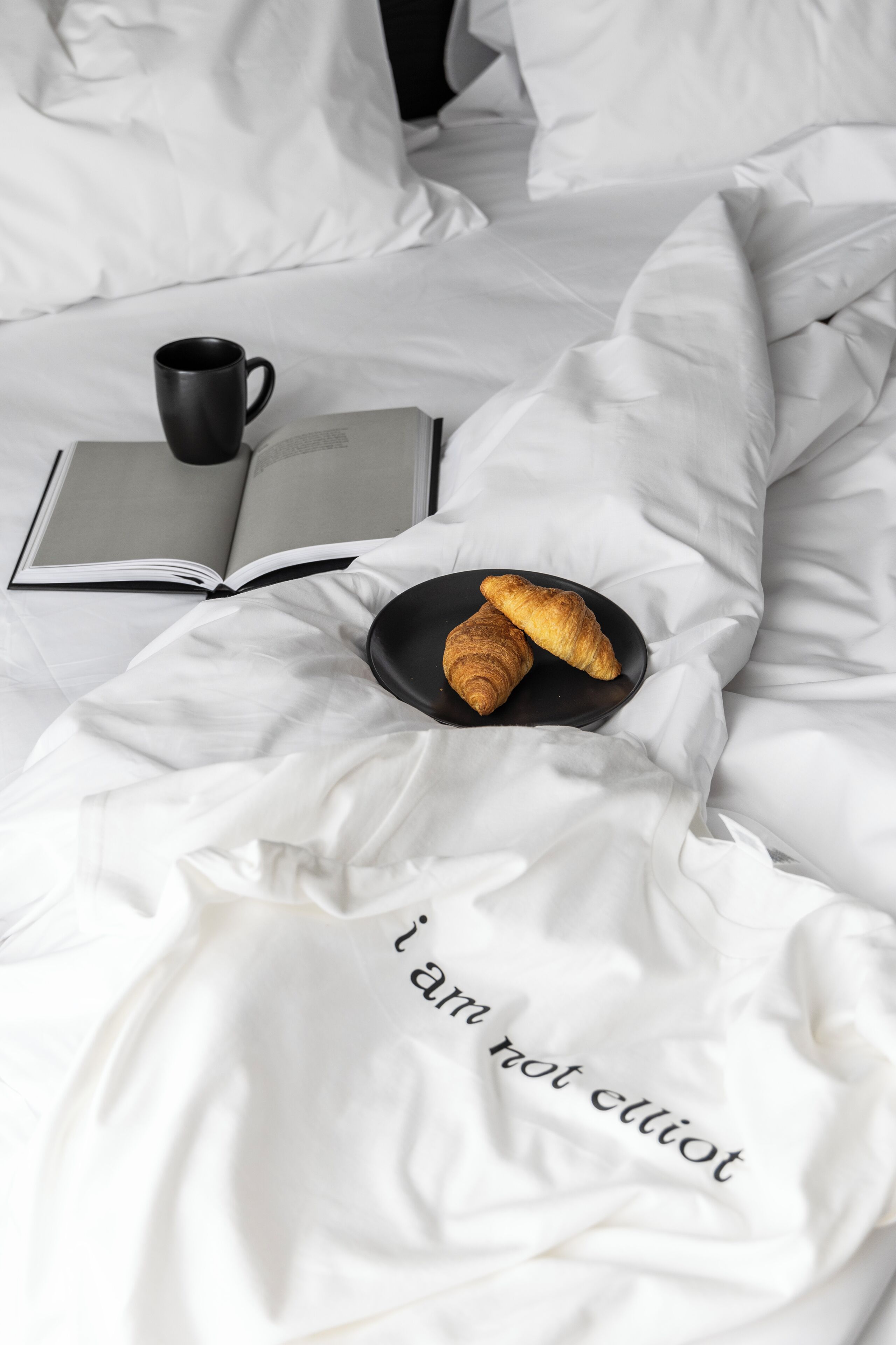 A bed with white linens, a black mug, an open book, and two croissants on a black plate.