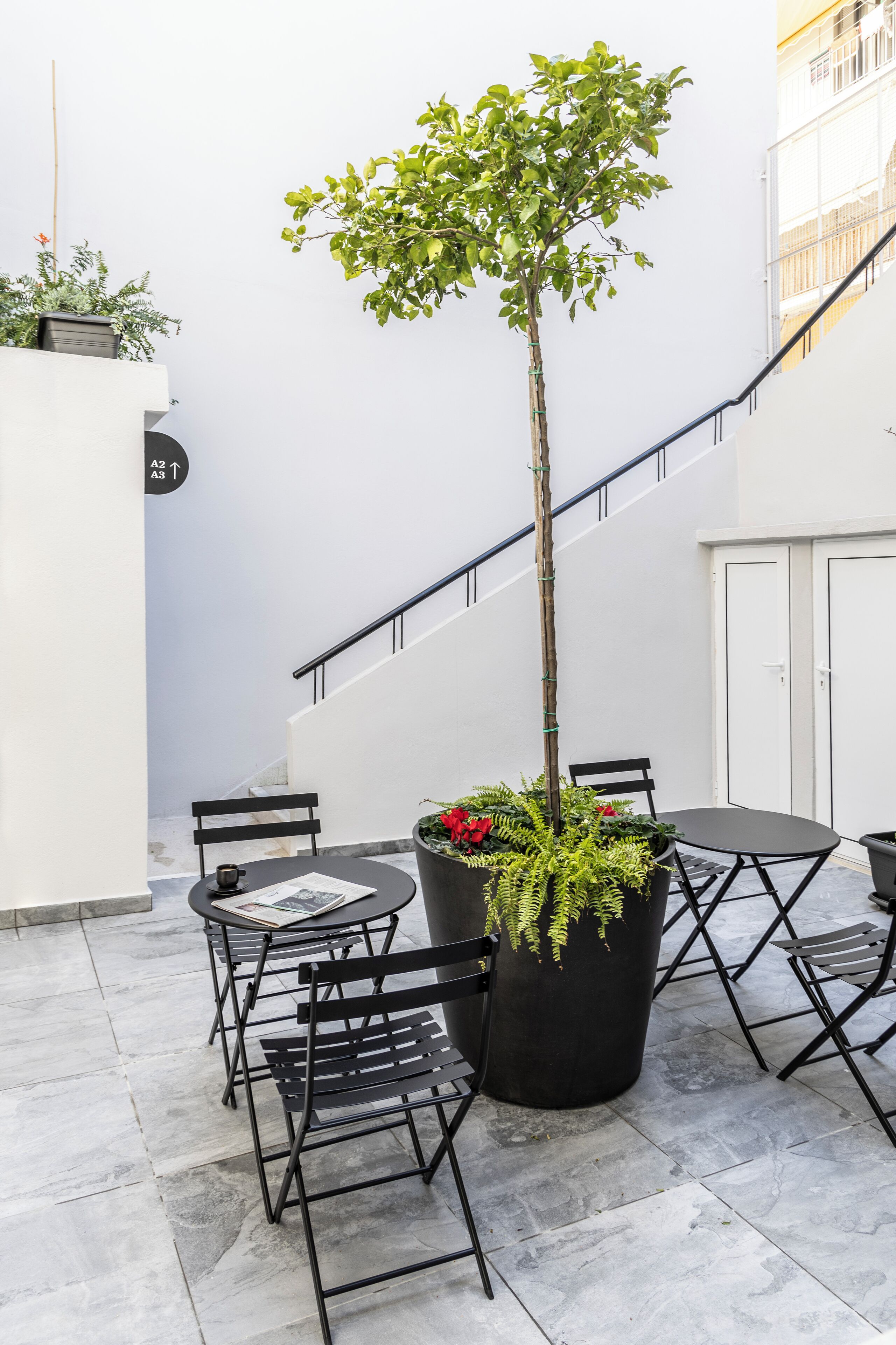 Una terraza con una mesa y sillas de metal negro, una maceta con flores rojas y una escalera.