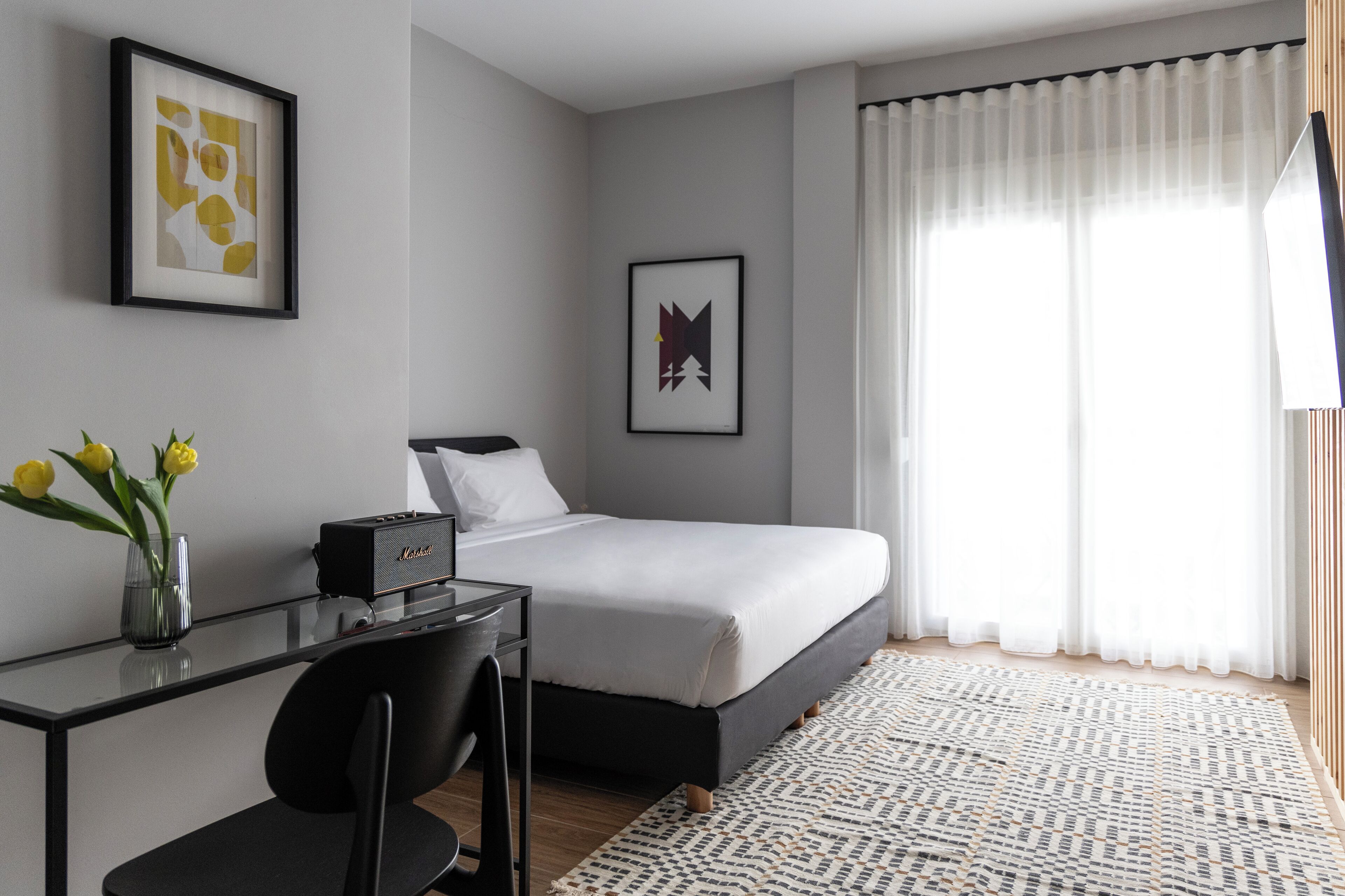 Una habitació d'hotel moderna amb un llit, escriptori, cadira i una finestra gran amb cortines.