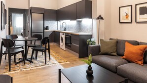 A modern kitchen with dark cabinetry, a built-in oven, and a sink. There is a dining area with a table and chairs, and a living area with a sofa and cushions. - Elliot Petralona, Athens (Athens)