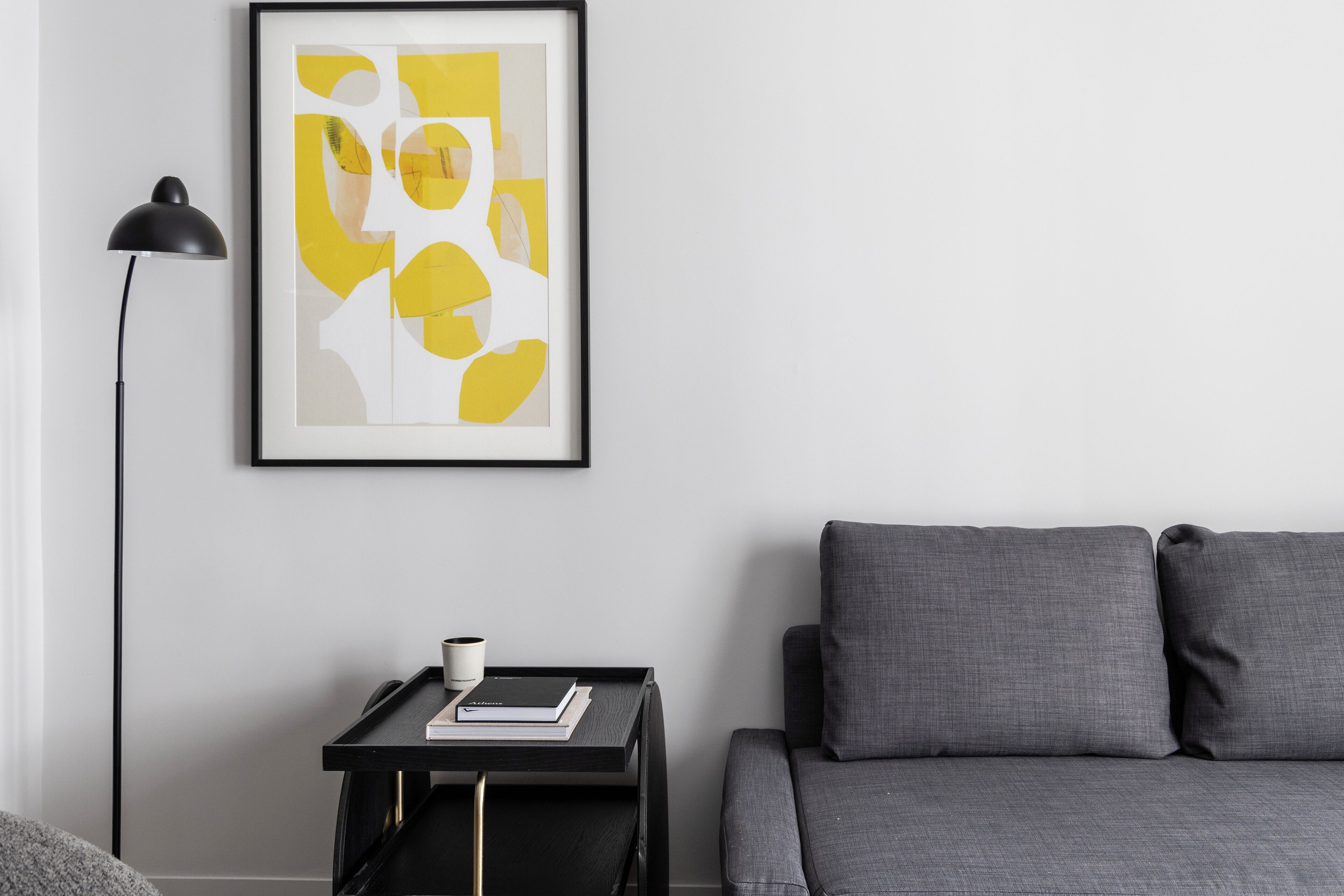 A modern living room with a grey sofa, a black side table with a book and a cup, and a framed abstract art piece on the wall.