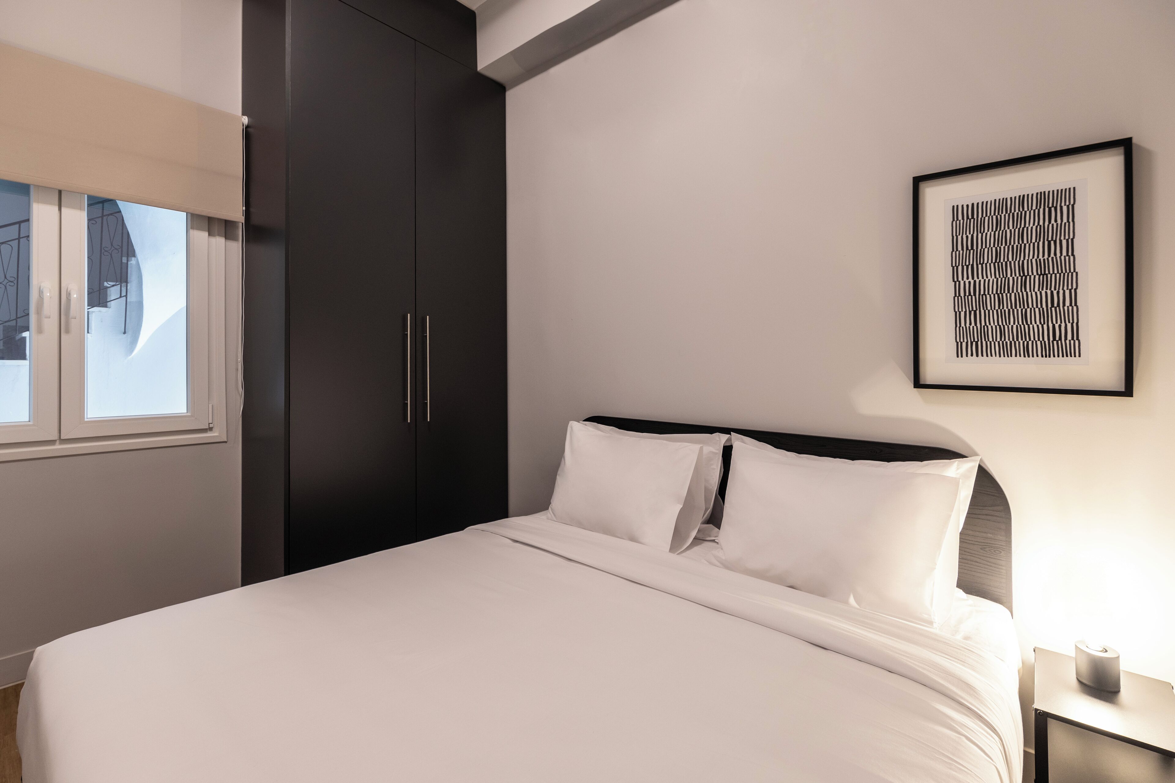A neatly made bed with white linens, a dark wardrobe, a framed abstract artwork on the wall, and a bedside table with a lamp.