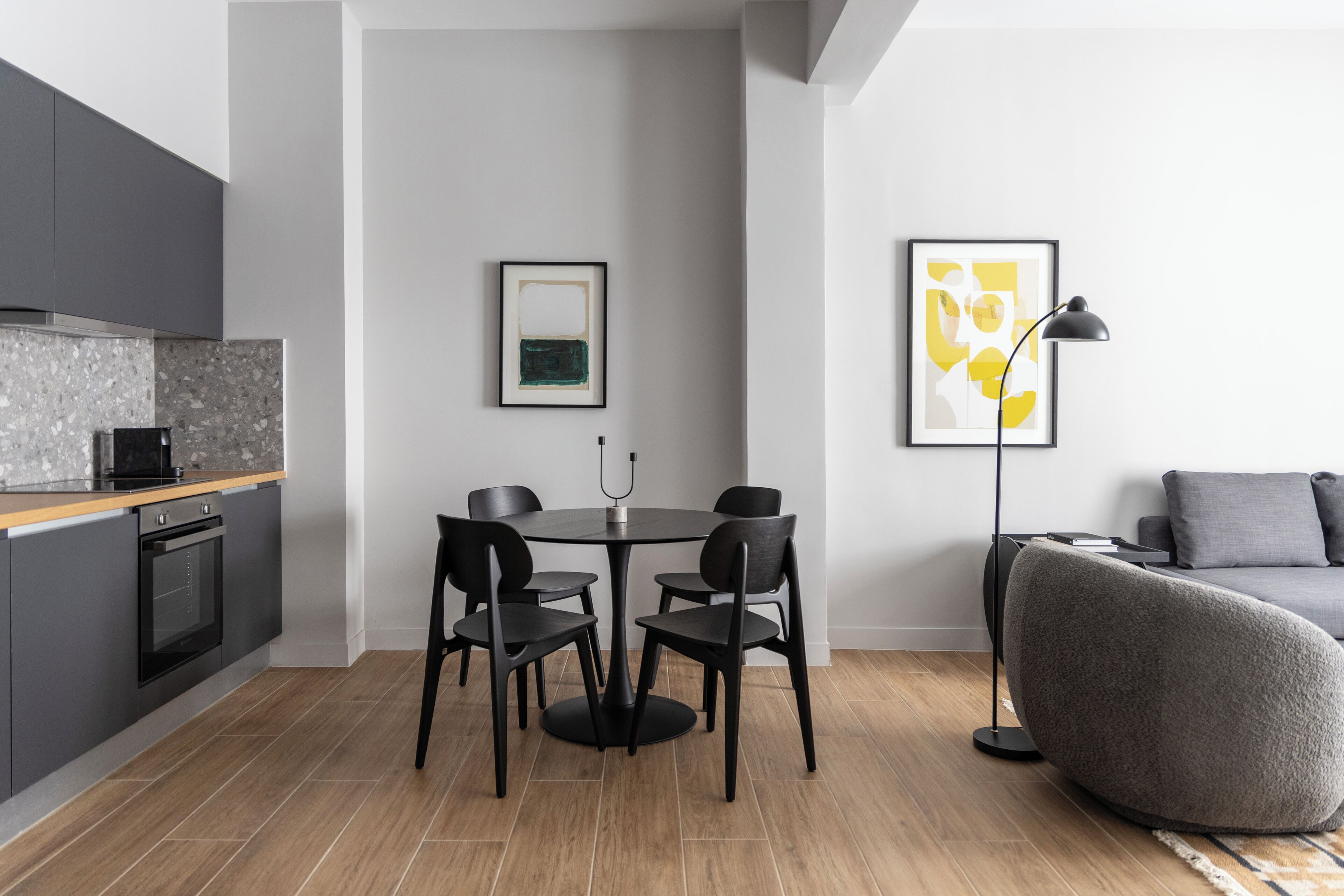 A modern kitchen with dark cabinetry, a built-in oven, and a dining area with a round table and four chairs. There is a framed abstract painting on the wall and a floor lamp beside a gray sofa.