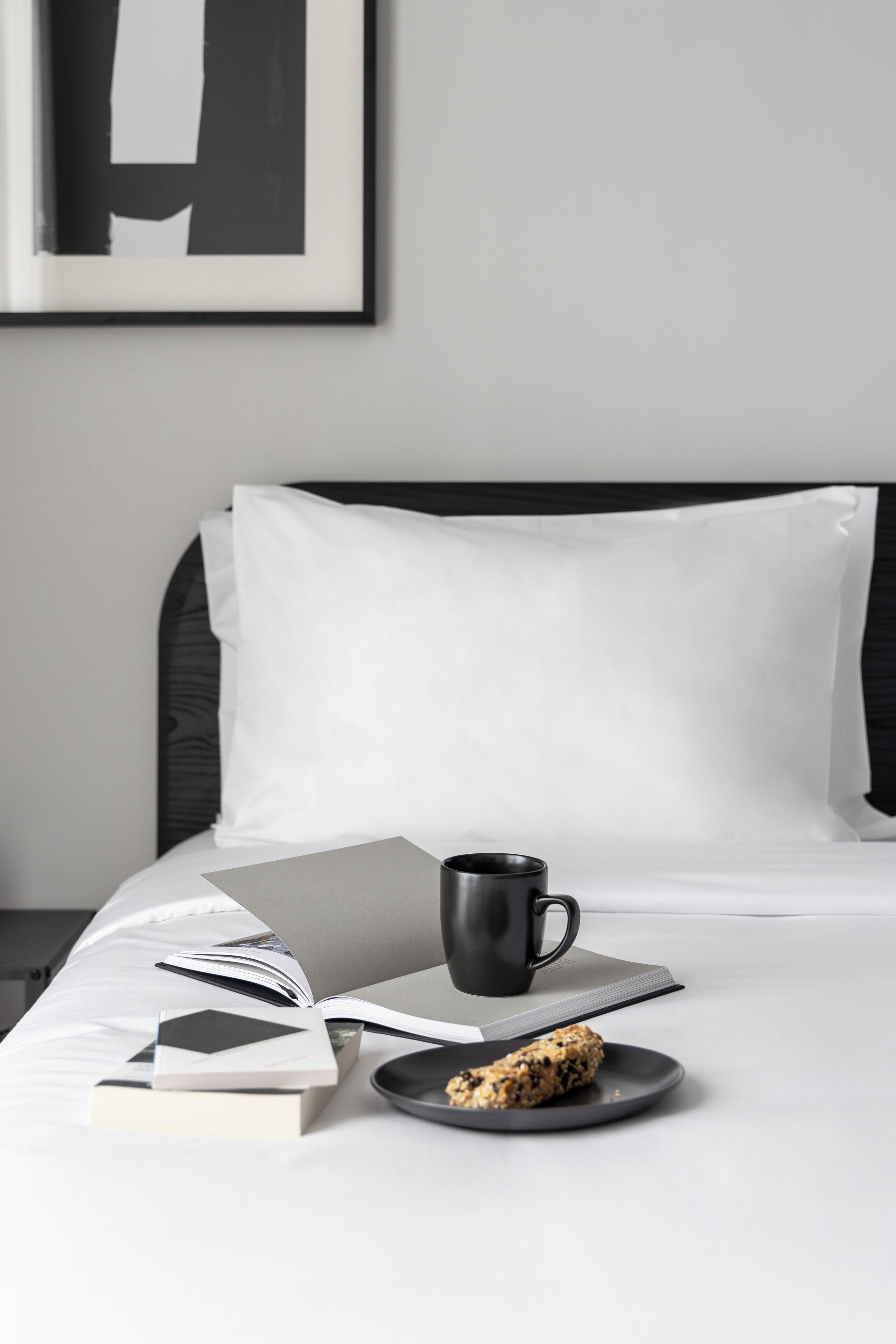 A neatly made bed with a black headboard, a white pillow, a black mug, a book, and a plate with a biscuit.