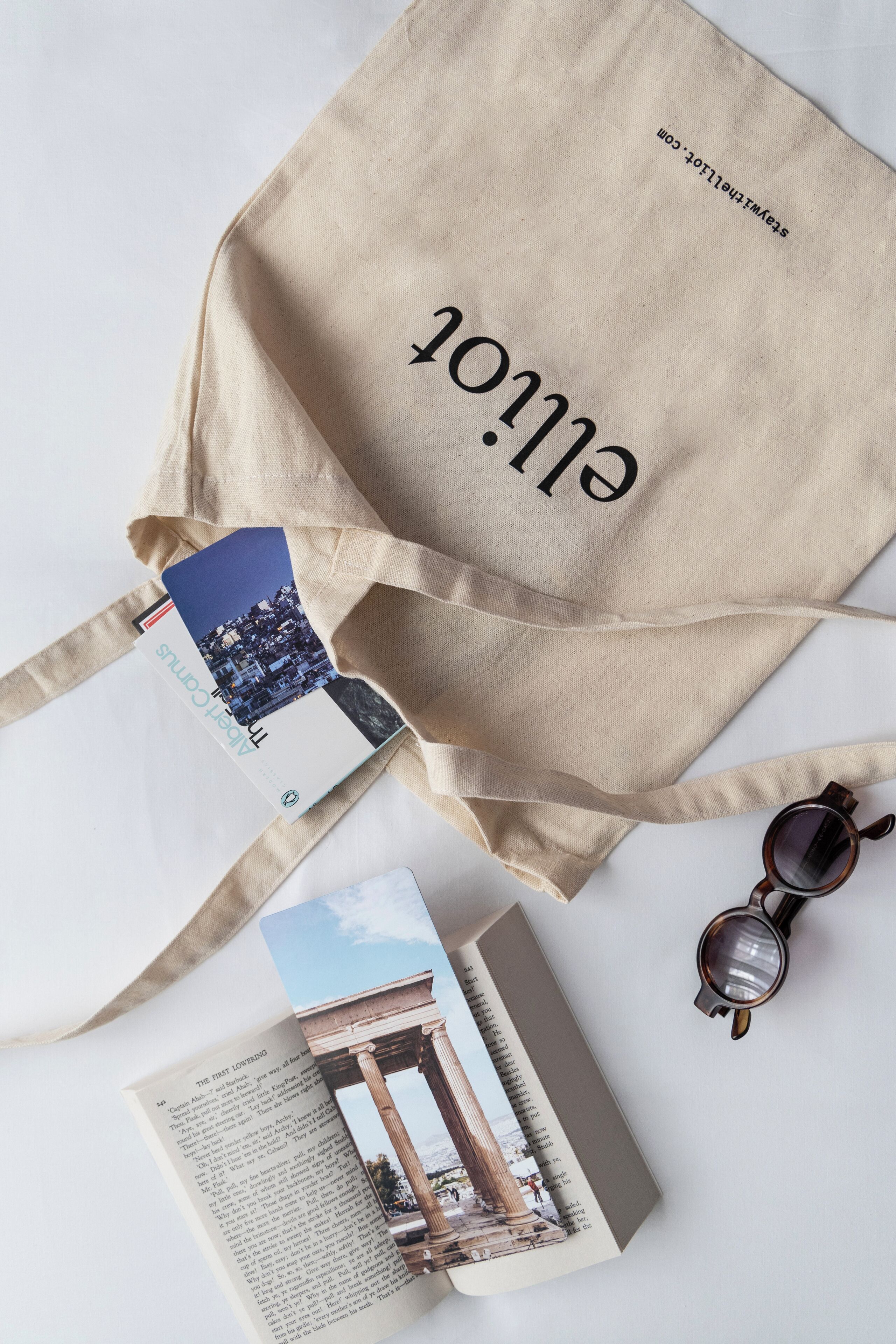 A beige tote bag with "ELOLA" printed on it, a pair of sunglasses, and a book with a picture of a building on the cover.