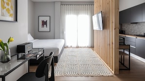 A modern hotel room with a bed, a desk with a computer, a TV, and a kitchen area. - Elliot Petralona, Athens (Athens)